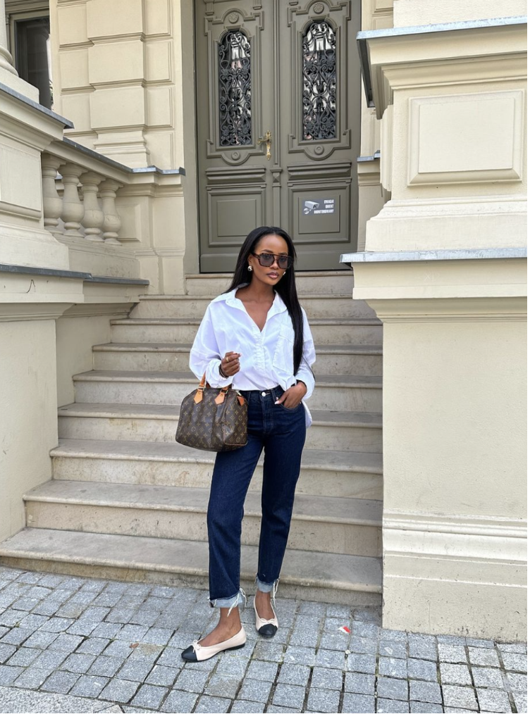 A woman wearing a white button-down shirt and light blue jeans paired with black Mary Jane flats, a classic ballet flats for women outfit idea.