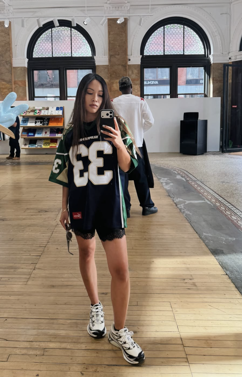 A woman styling an oversized black and green sports jersey as a dress over black lace-trimmed bike shorts, paired with chunky black and white sneakers.