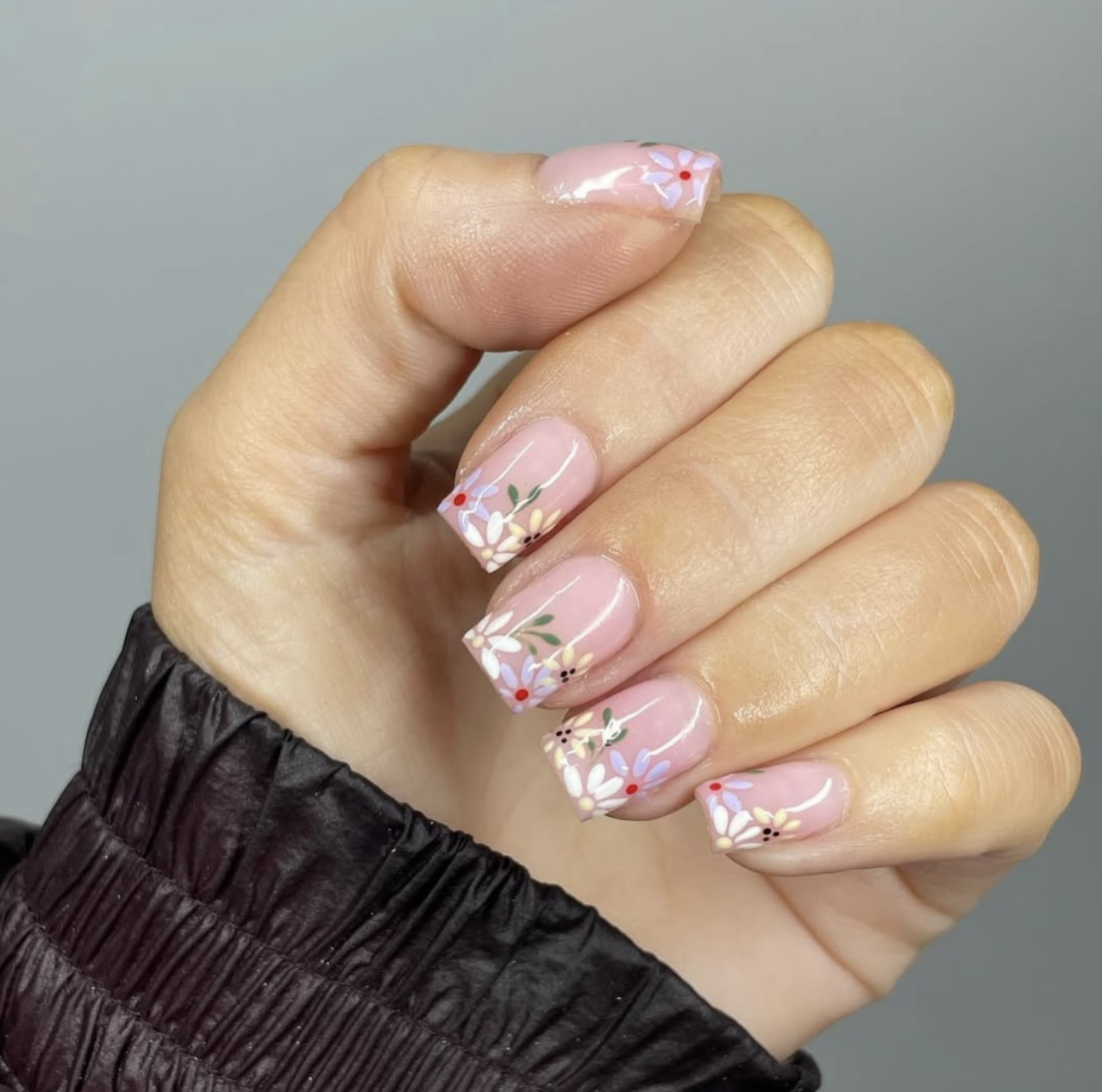 A close-up of a hand with short square-shaped nude gel nails featuring tiny, minimalist hand-painted blue and pink wildflowers.