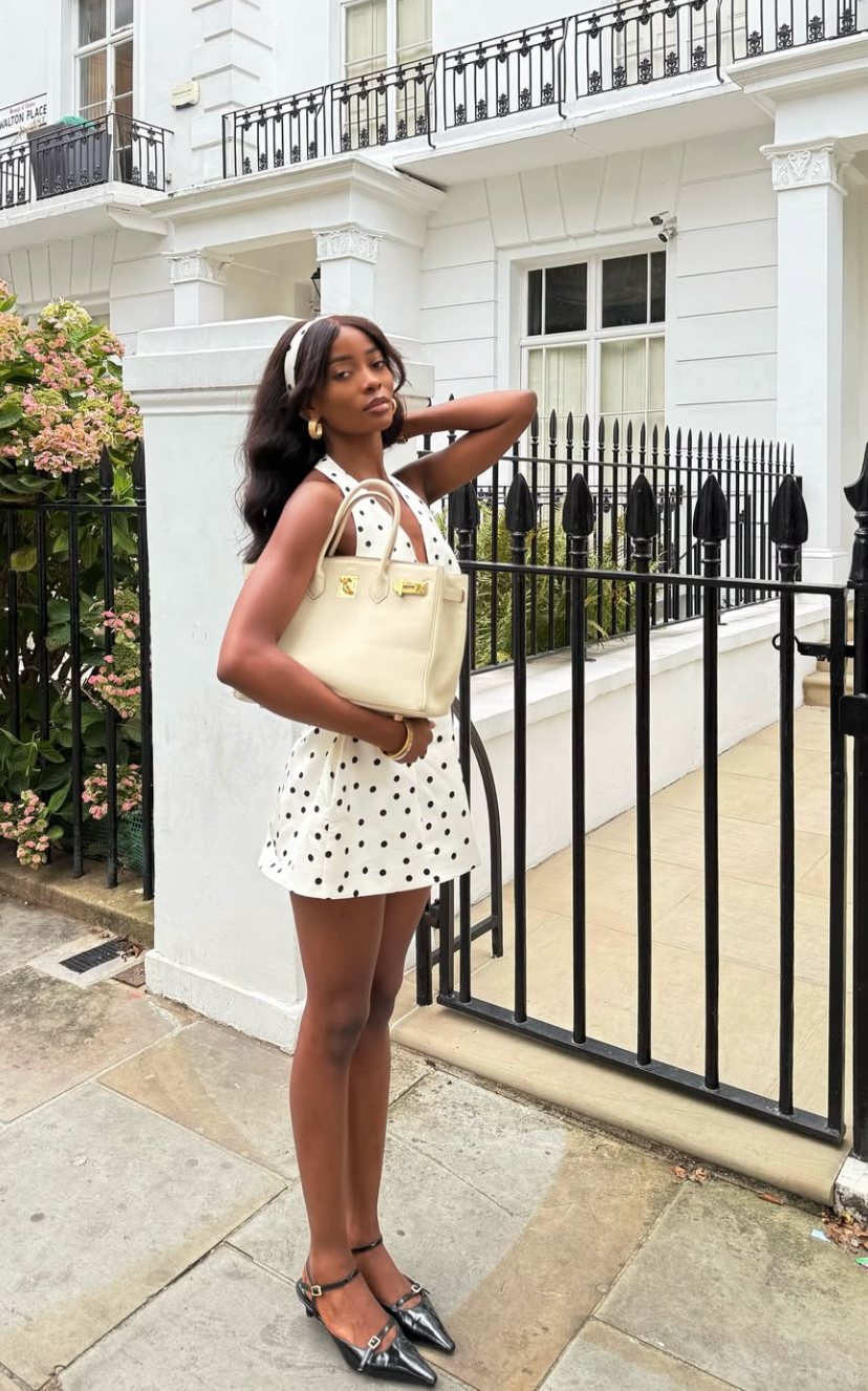 A woman wearing a white halter-neck mini dress with small black polka dots, paired with black pointed-toe slingback flats and a cream-colored handbag.