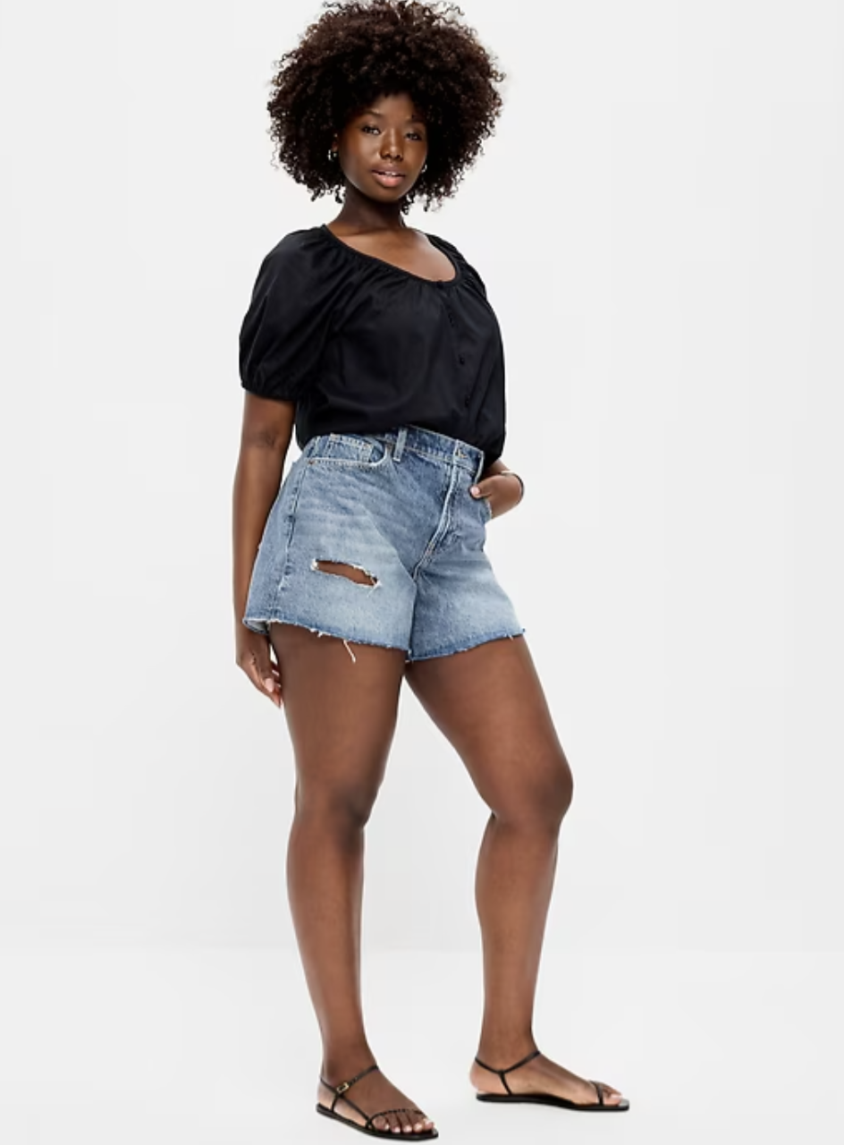 A woman with fuller legs wearing light-wash Old Navy Curvy high-waisted baggy denim cut-off shorts with a 5-inch inseam, styled with a black puff-sleeve top and black sandals.