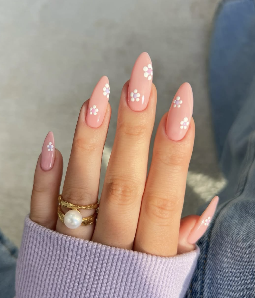 Long almond-shaped nails with a glossy pale pink base, decorated with minimalist hand-painted white and purple daisy flower accents, highlighting cute June nail ideas.