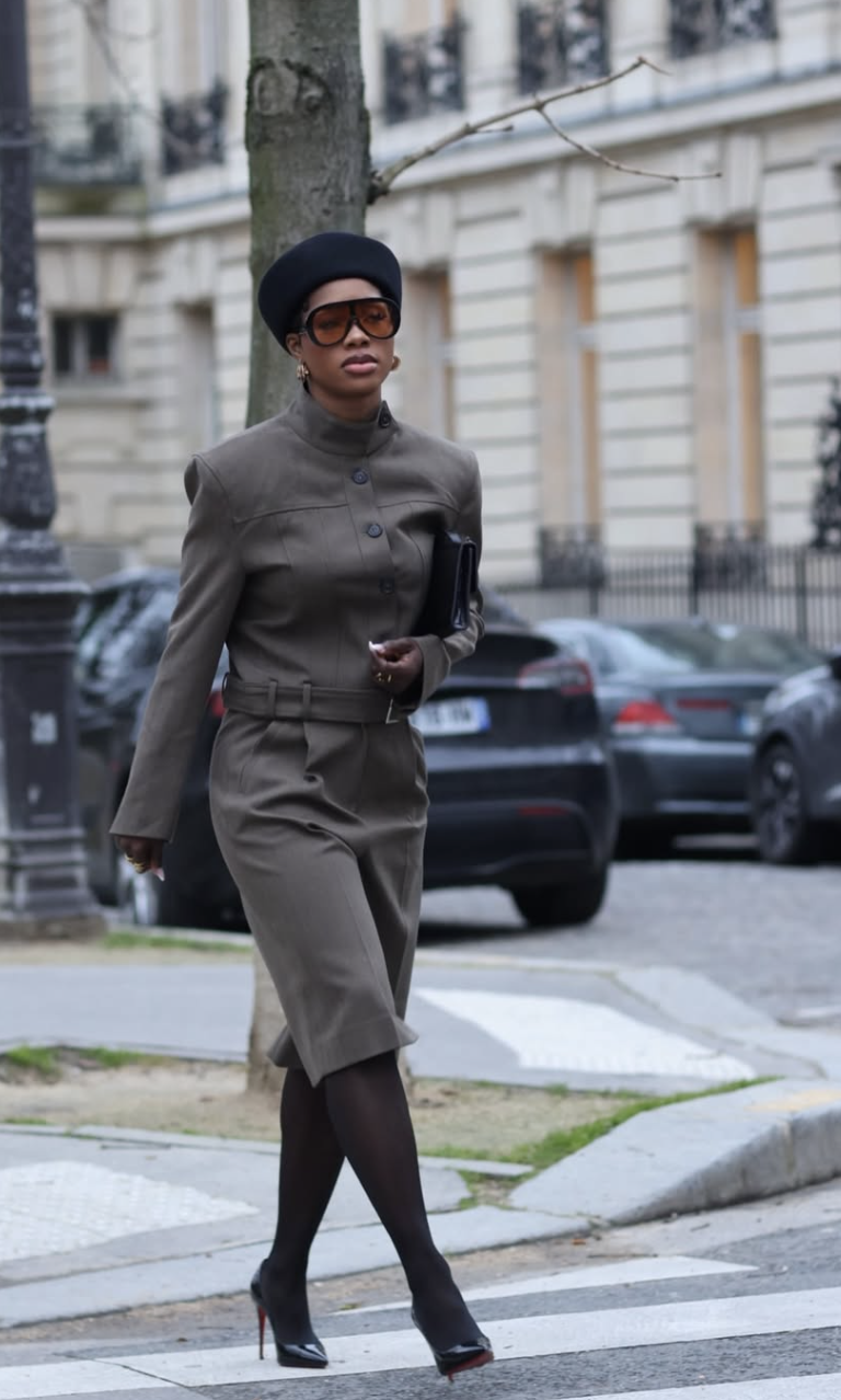 A woman wearing a black beret, large orange sunglasses, a tailored olive green high-neck jacket with a belt, and knee-high boots while walking outside.