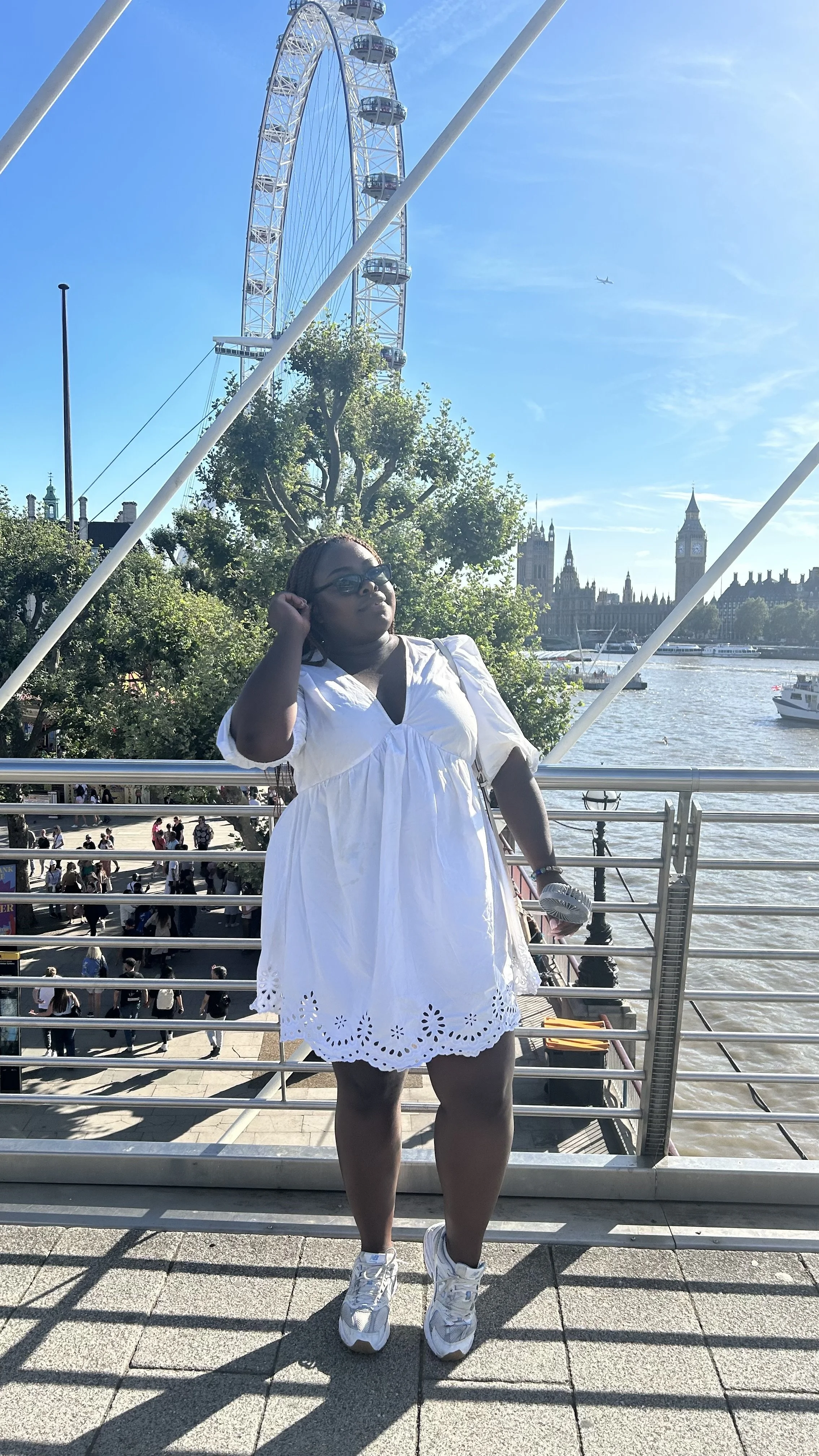 A woman wearing a white short-sleeved mini dress and white sneakers stands on a bridge with the London Eye in the background.