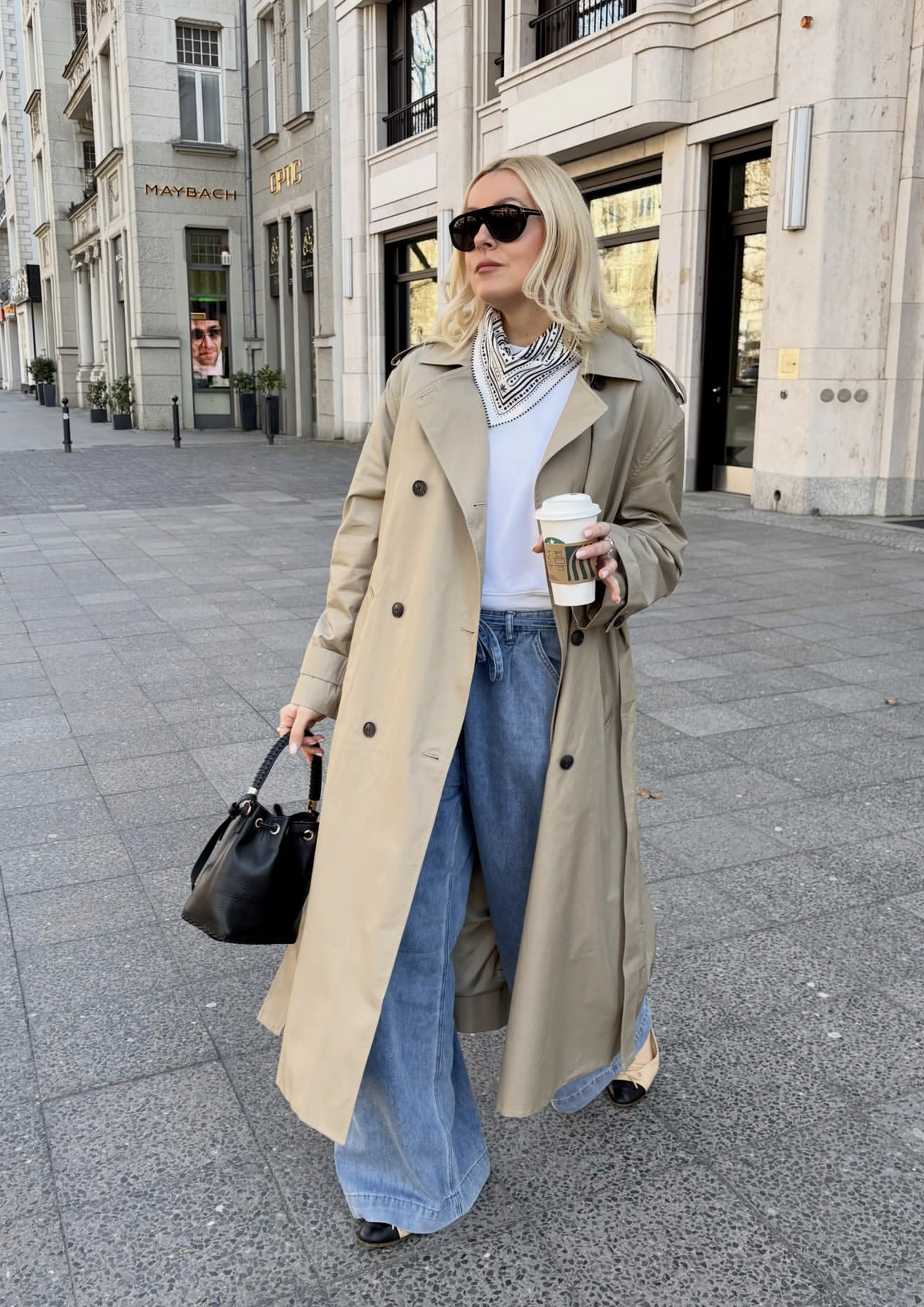 A woman walking down a city street wearing a long tan trench coat over a white t-shirt and light-wash wide-leg jeans, accessorized with a patterned neck scarf and a black bucket bag.