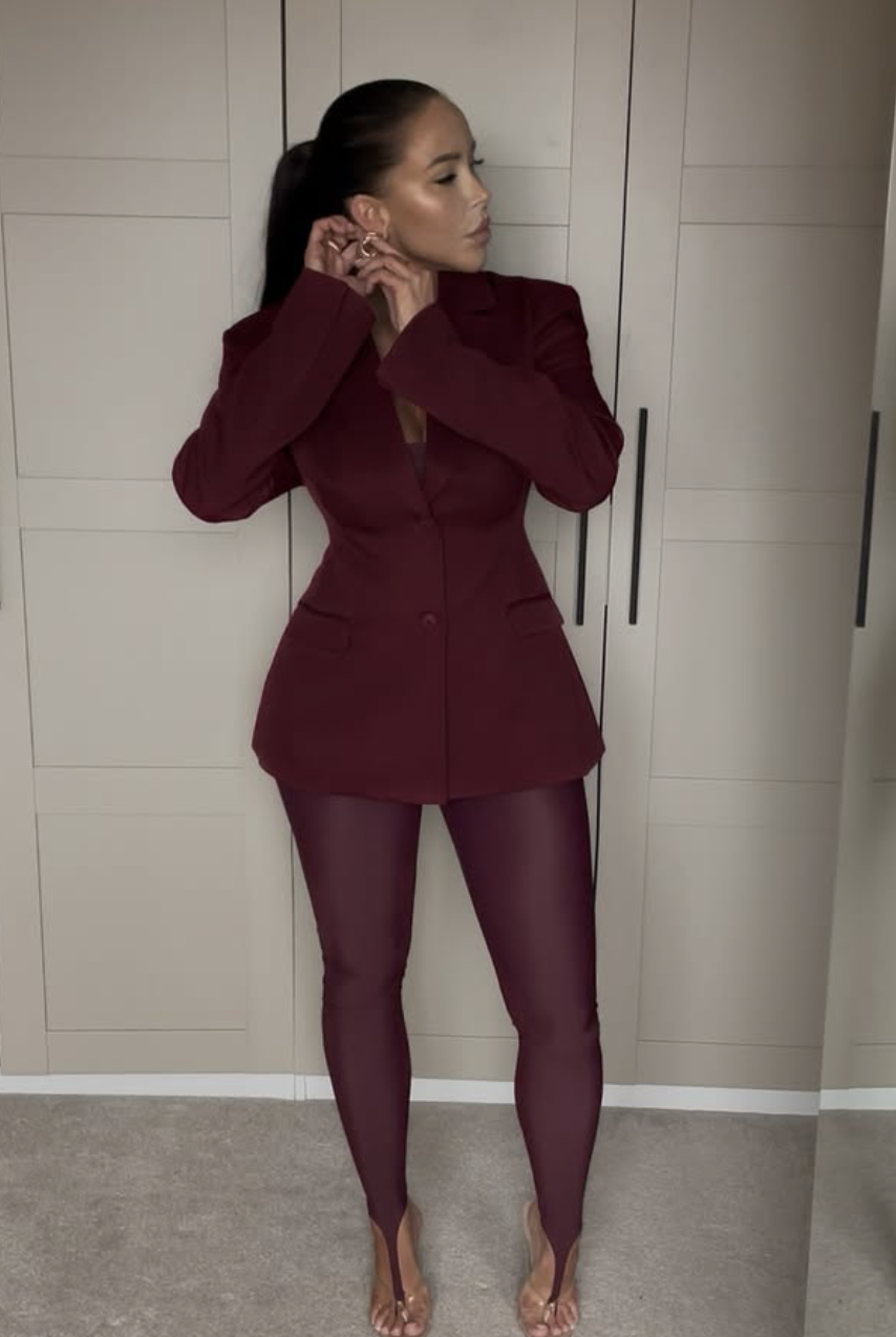 Woman wearing a monochrome burgundy outfit featuring a cinched waist blazer, matching stirrup leggings, and pointed-toe heels.