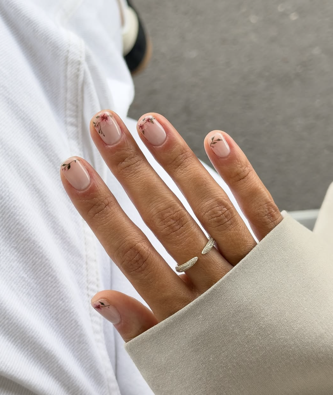 A close-up of short, natural nails featuring delicate hand-painted red and green floral accents with a clear negative space base.