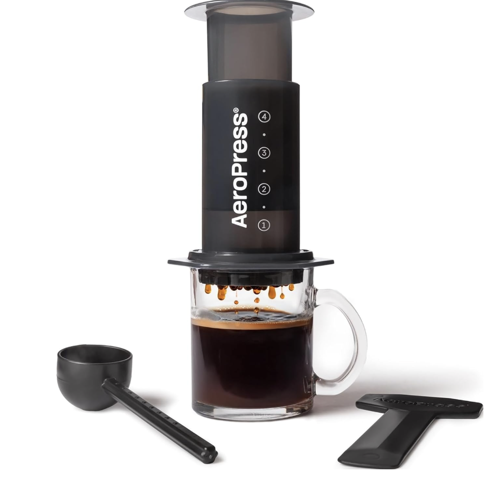 AeroPress Original coffee maker resting on a clear glass mug while brewing coffee, displayed alongside a black scoop.