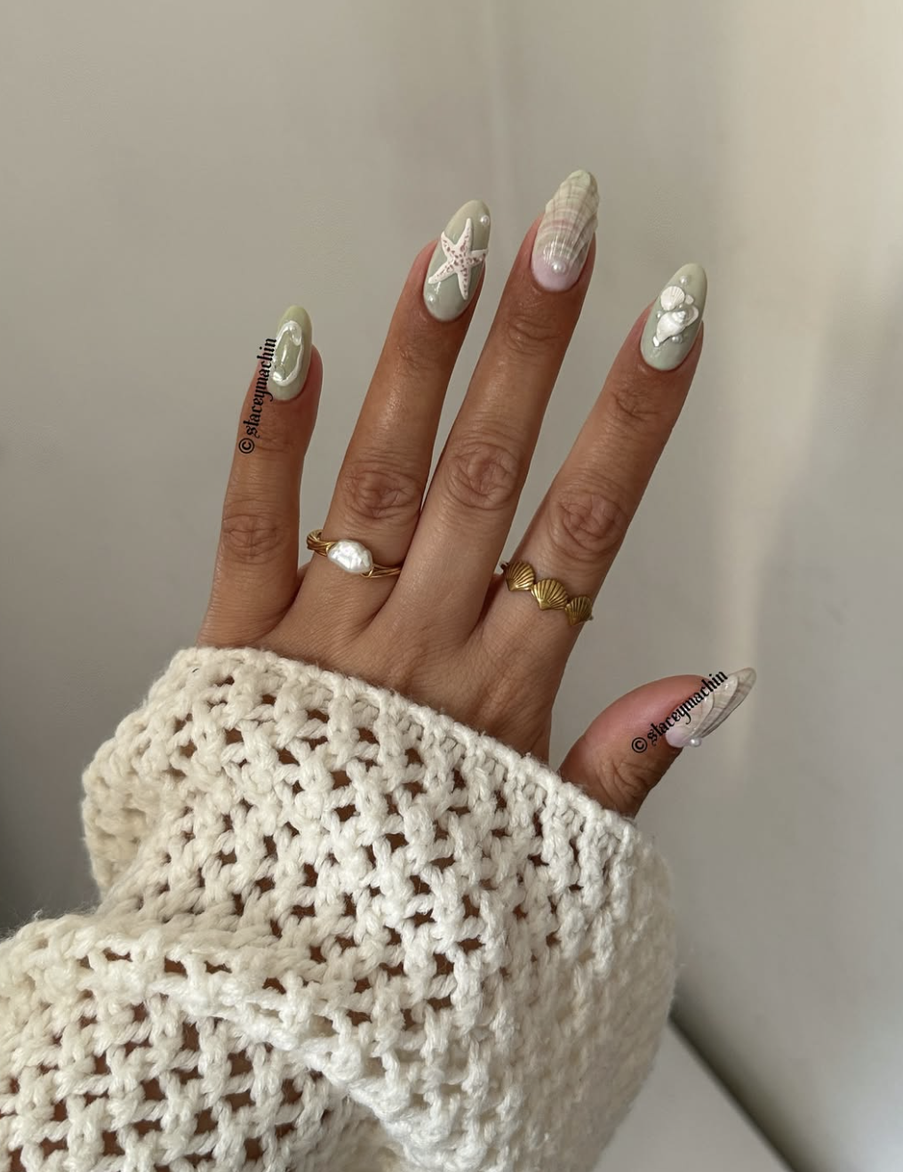 A close-up of long, almond-shaped nails featuring a muted sage-green base with 3D ocean-themed artwork, including a white starfish, a realistic textured seashell, and tiny pearl embellishments, paired with a white knit sleeve.