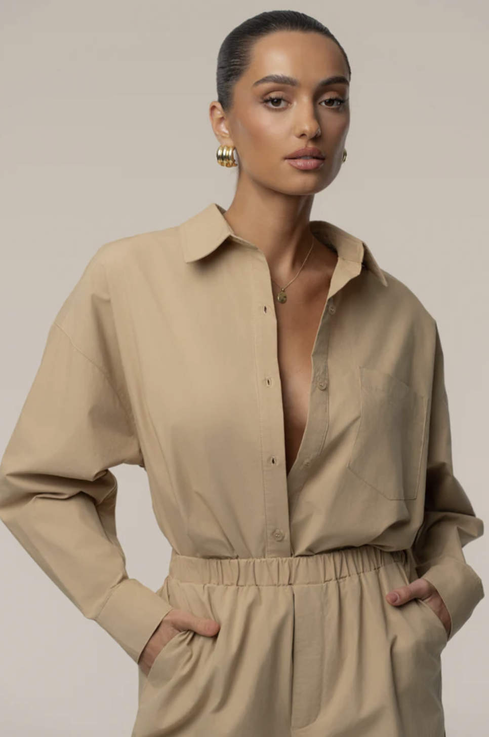 A product shot of a woman wearing a beige Leon poplin shirt tucked into matching tailored trousers. This illustrates a monochromatic tailored suit set that can be layered with a white t-shirt for a modern street-style look.