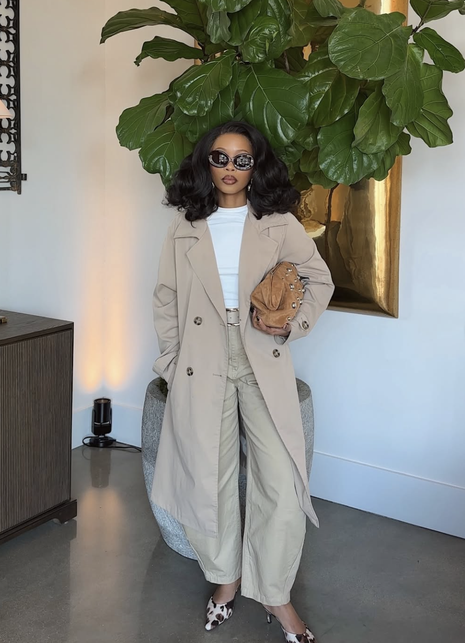 A woman wearing a tan maxi trench coat over a white bodysuit and beige wide-leg trousers, accessorized with a brown woven clutch and animal print heels.