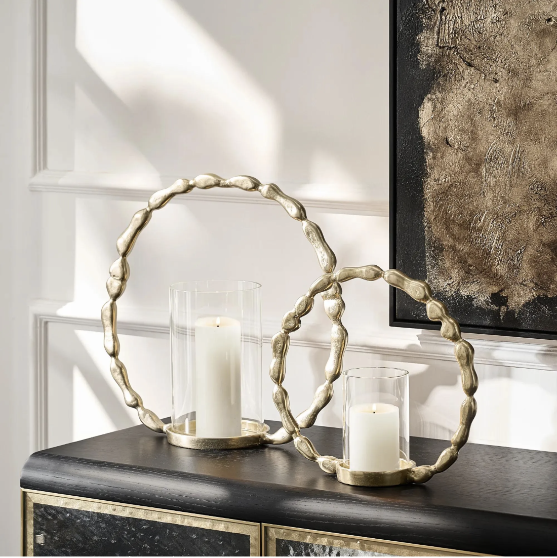 Two gold organic ring hurricane candle holders with white pillar candles on a black console beneath abstract art.