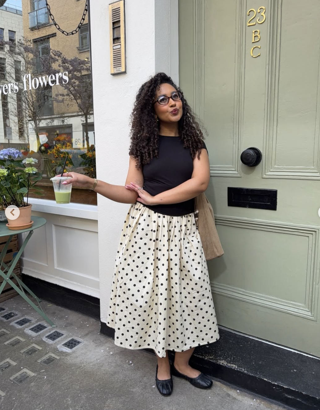 A woman standing on a city sidewalk wearing a black short-sleeved top and a cream-colored midi skirt with black polka dots and a wide white lace trim at the hem.