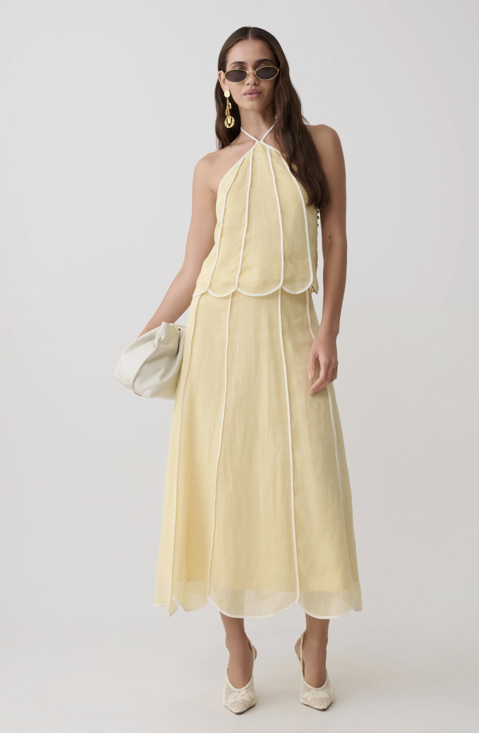 A woman wearing a pale yellow halter top with white scalloped trim and contrast piping, accessorized with large gold statement earrings and dark oval sunglasses.