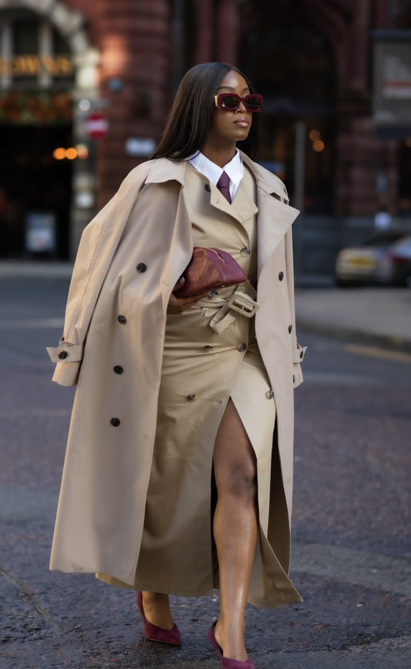 A woman styling a tan trench coat dress with a high-waisted belt and a button-down shirt to hide a long torso and create the illusion of longer legs.