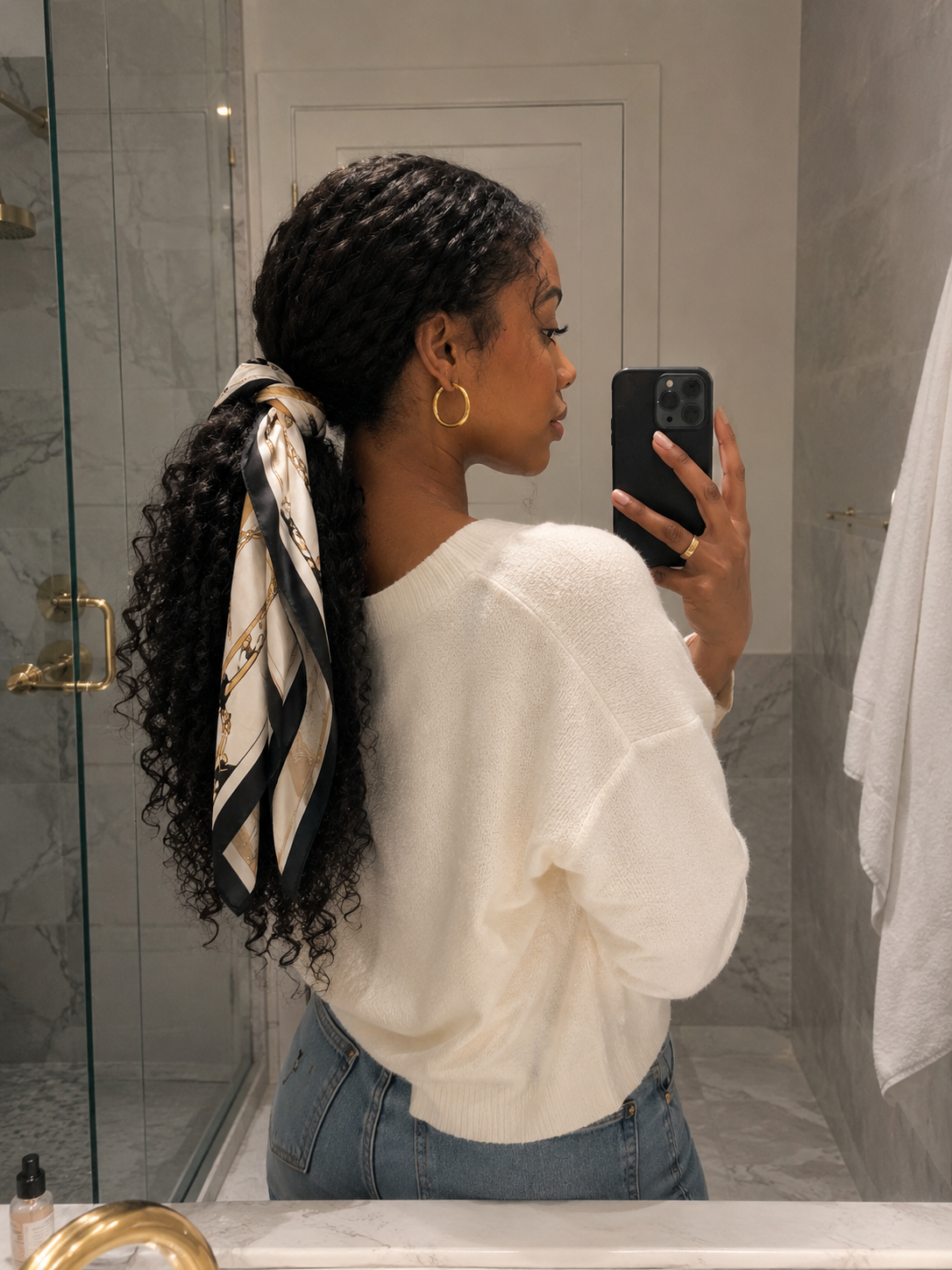 A woman from behind showing how to style a printed silk scarf tied around a loose low ponytail in dark curly hair.