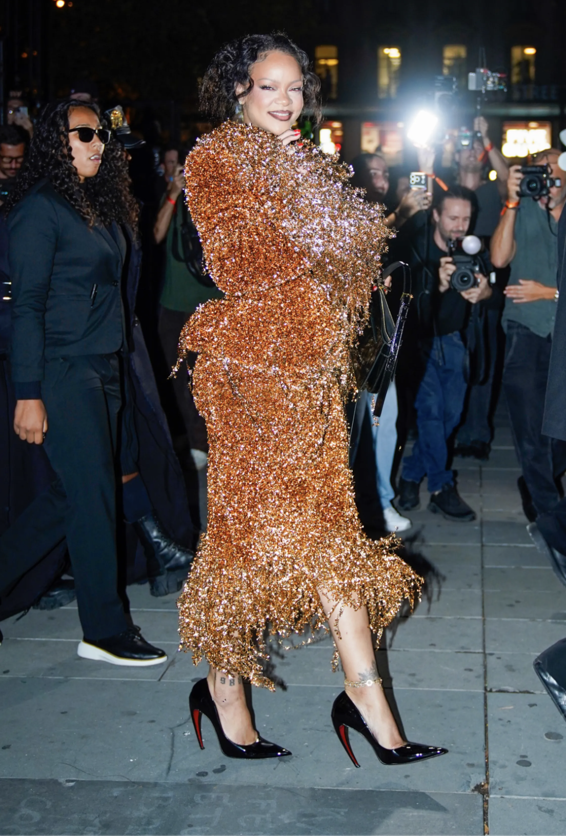 Rihanna wears a bronze metallic-thread skirt suit and black curved-heel pumps at the Matières Fécales Spring 2027 show in Paris.