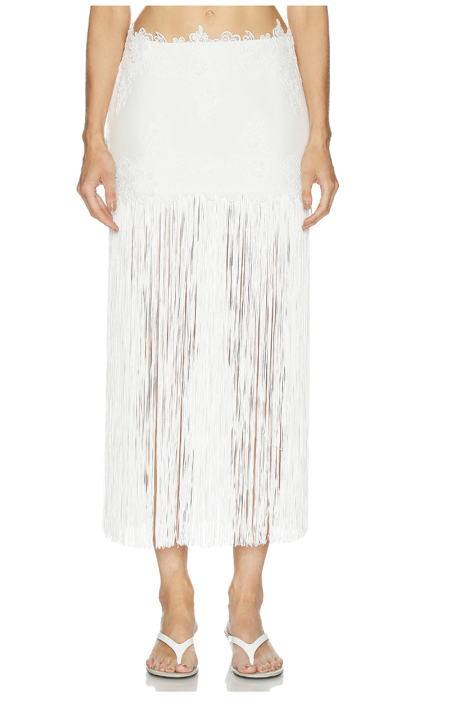 A close-up view of a woman wearing the white Retrofete Amaris fringe midi skirt paired with white thong sandals.
