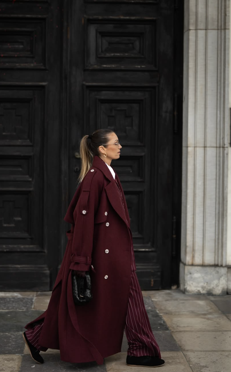 A woman in profile wearing a long burgundy trench-style coat with large silver buttons and shoulder epaulettes standing in front of a dark door.