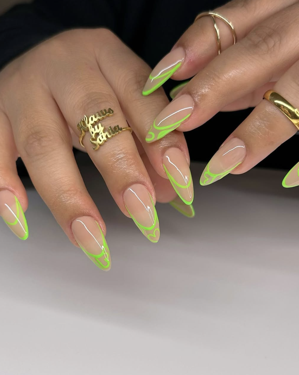 A close-up of long, almond-shaped nails with a sheer nude base, featuring a vibrant neon lime green abstract French tip outline with delicate wavy line patterns, accessorized with a customized name ring.