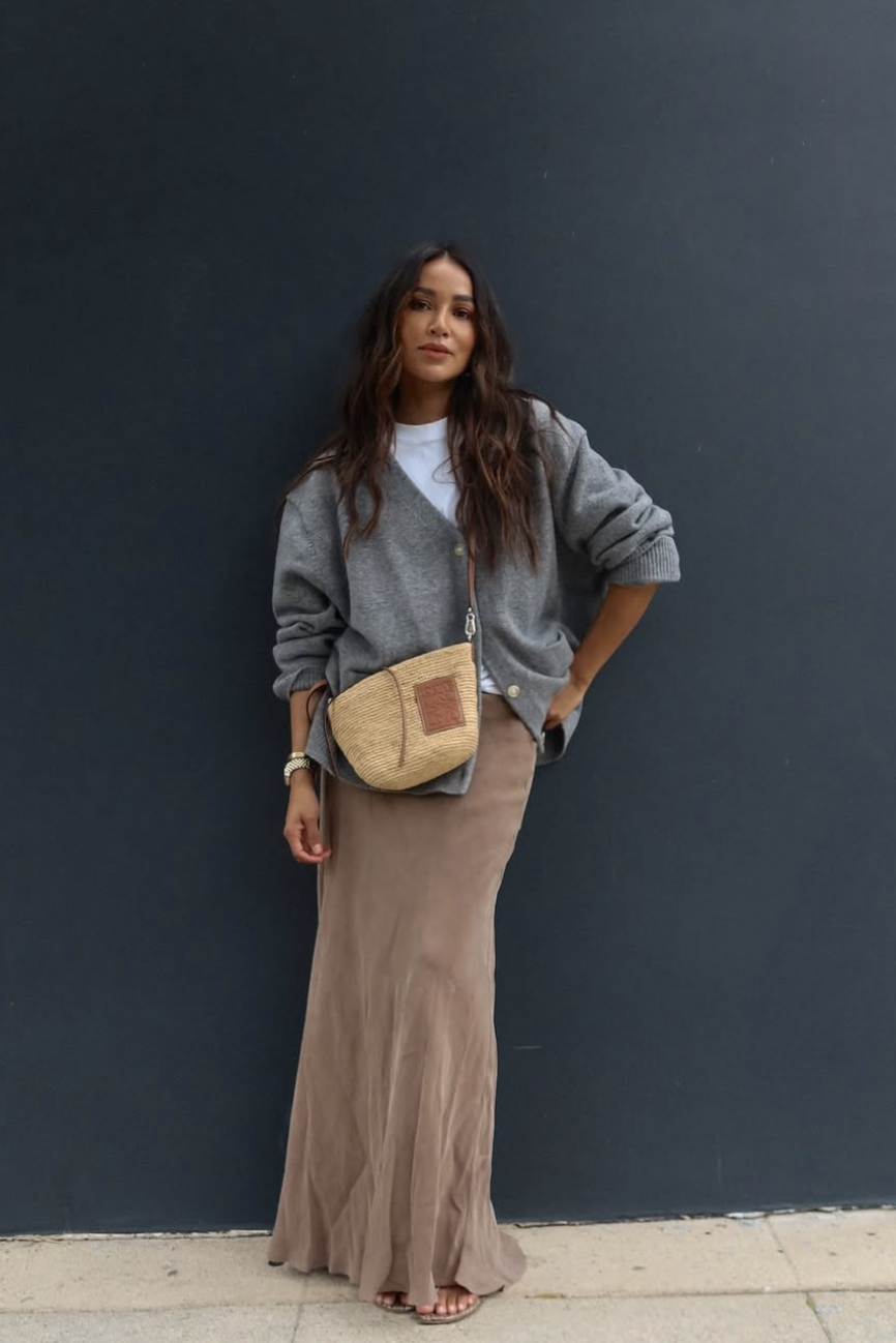 A woman wearing a brown maxi skirt, a white t-shirt, and an oversized grey cardigan, carrying a straw shoulder bag.