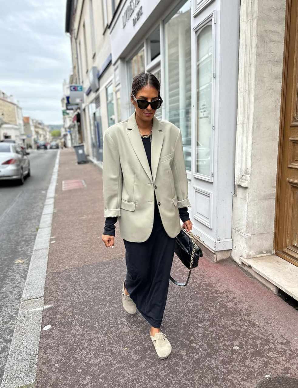 A woman walking down a city street styling an oversized sage green blazer over a black maxi dress, paired with light gray suede clogs and black sunglasses.