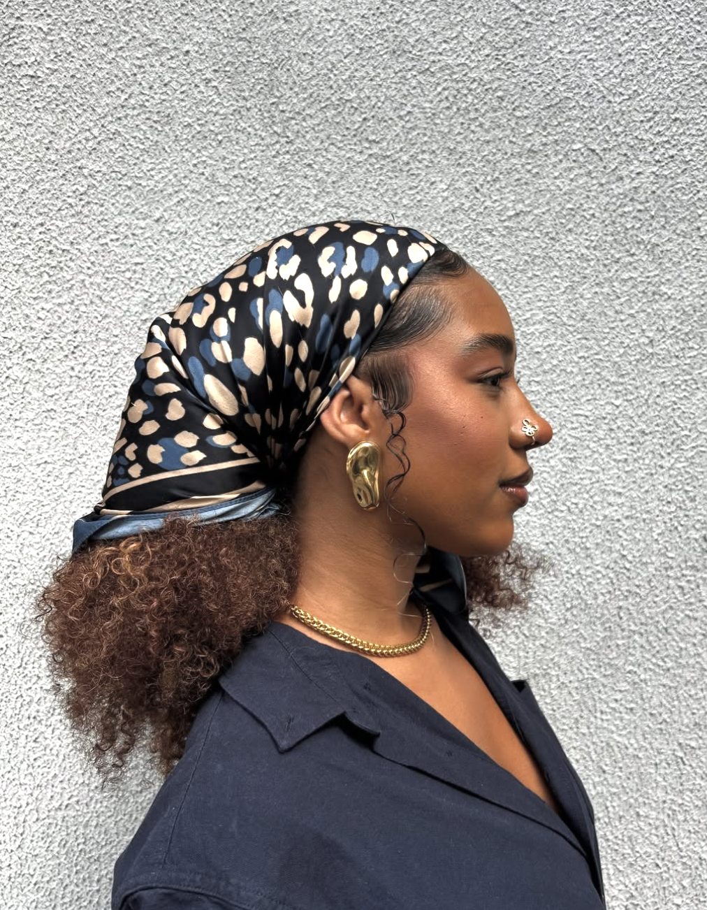 A close-up side profile of a woman with curly hair styled in an updo, wearing a black and tan leopard print silk scarf wrapped as a wide headband, paired with a chunky gold statement earring and chain necklace.