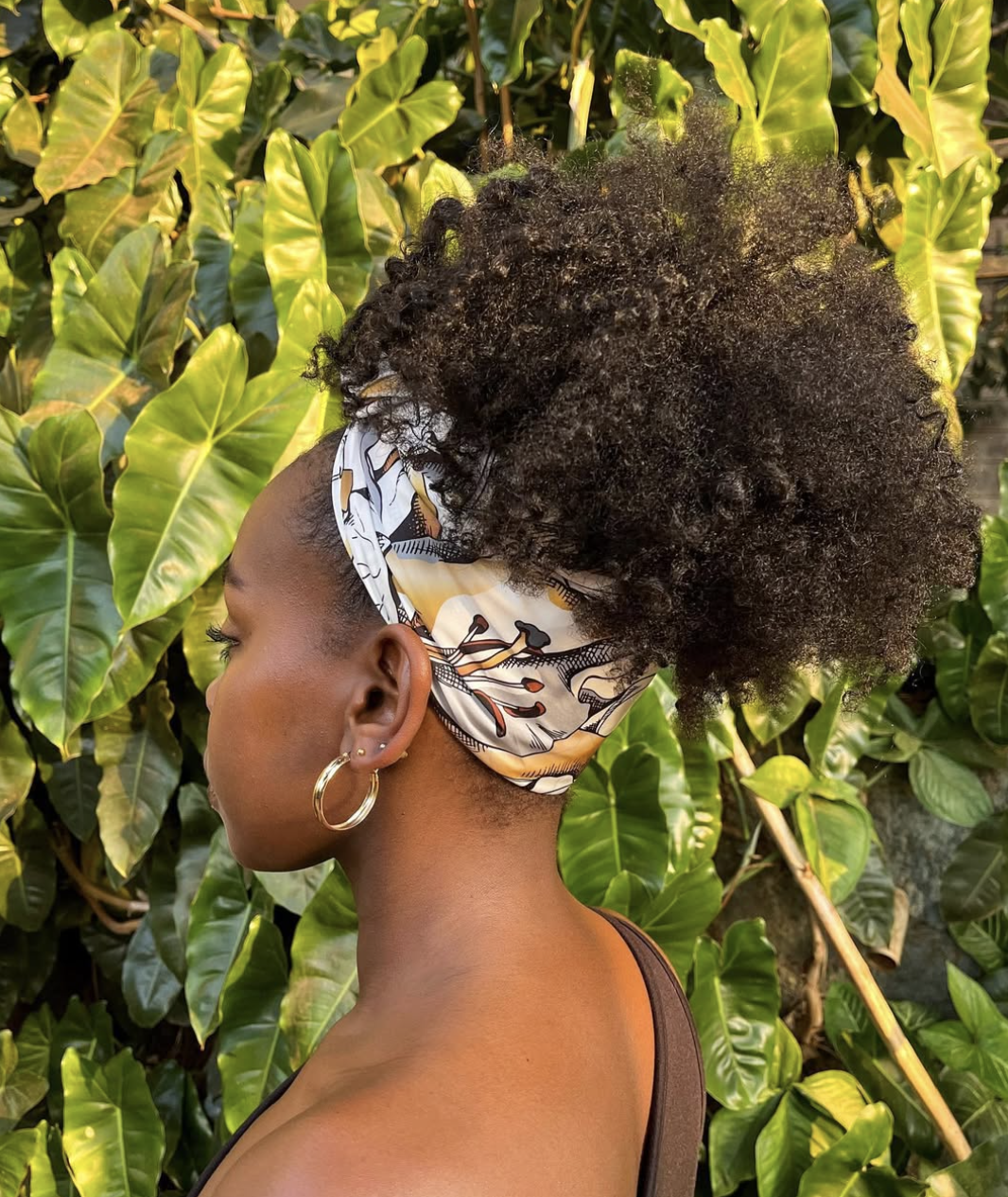 A woman seen from the side showing a patterned silk scarf wrapped around the base of a high curly pineapple puff updo with gold hoop earrings.