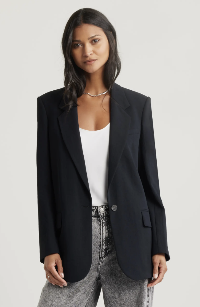 A woman wearing the Open Edit "The Curator" relaxed blazer in black over a white top and grey acid-wash jeans—a staple layering piece for a modern hybrid work capsule wardrobe.