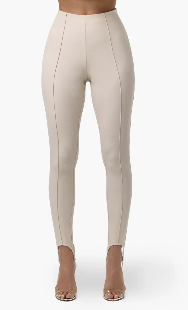 A woman wearing tan Naked Wardrobe suiting stirrup leggings with a visible front seam, paired with transparent pointed-toe heels—a modern and comfortable staple for a hybrid work capsule wardrobe.