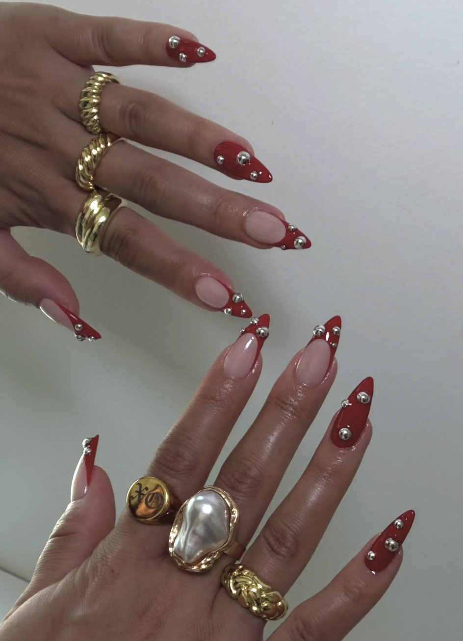 Long almond-shaped nails with a sheer nude base, vibrant red French tips, and varying sizes of 3D silver metallic studs scattered across the nail bed.