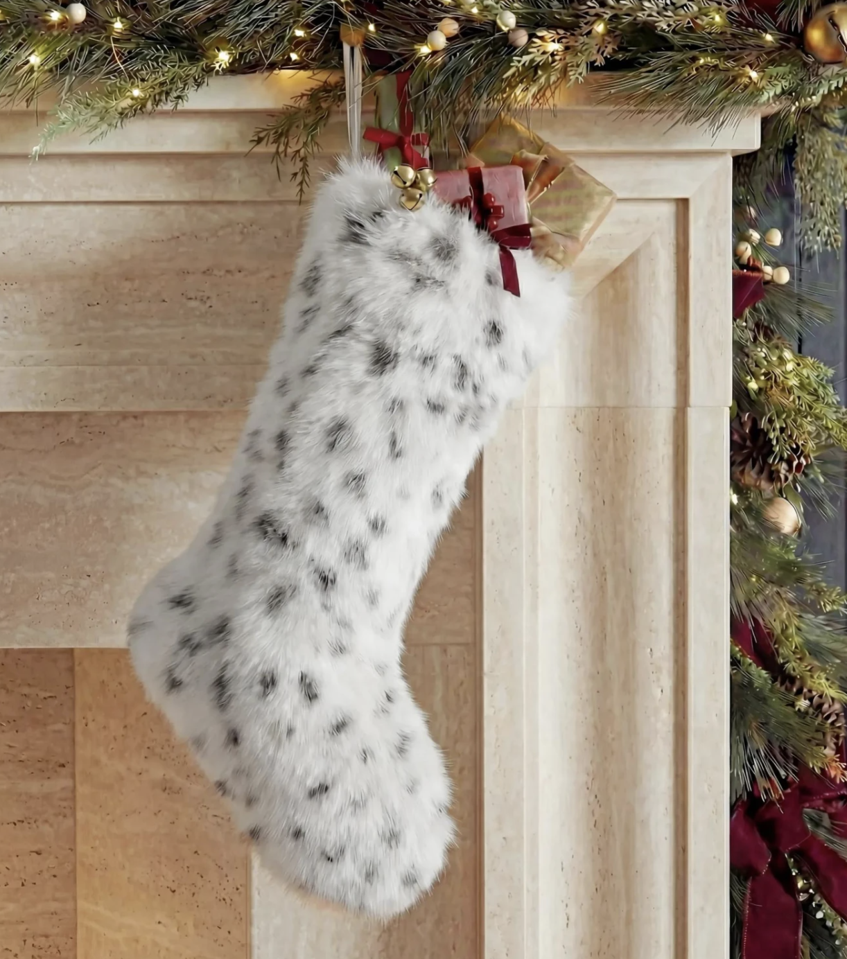 White faux fur snow leopard print Christmas stocking hanging from a stone mantel trimmed with lit garland and wrapped gifts.
