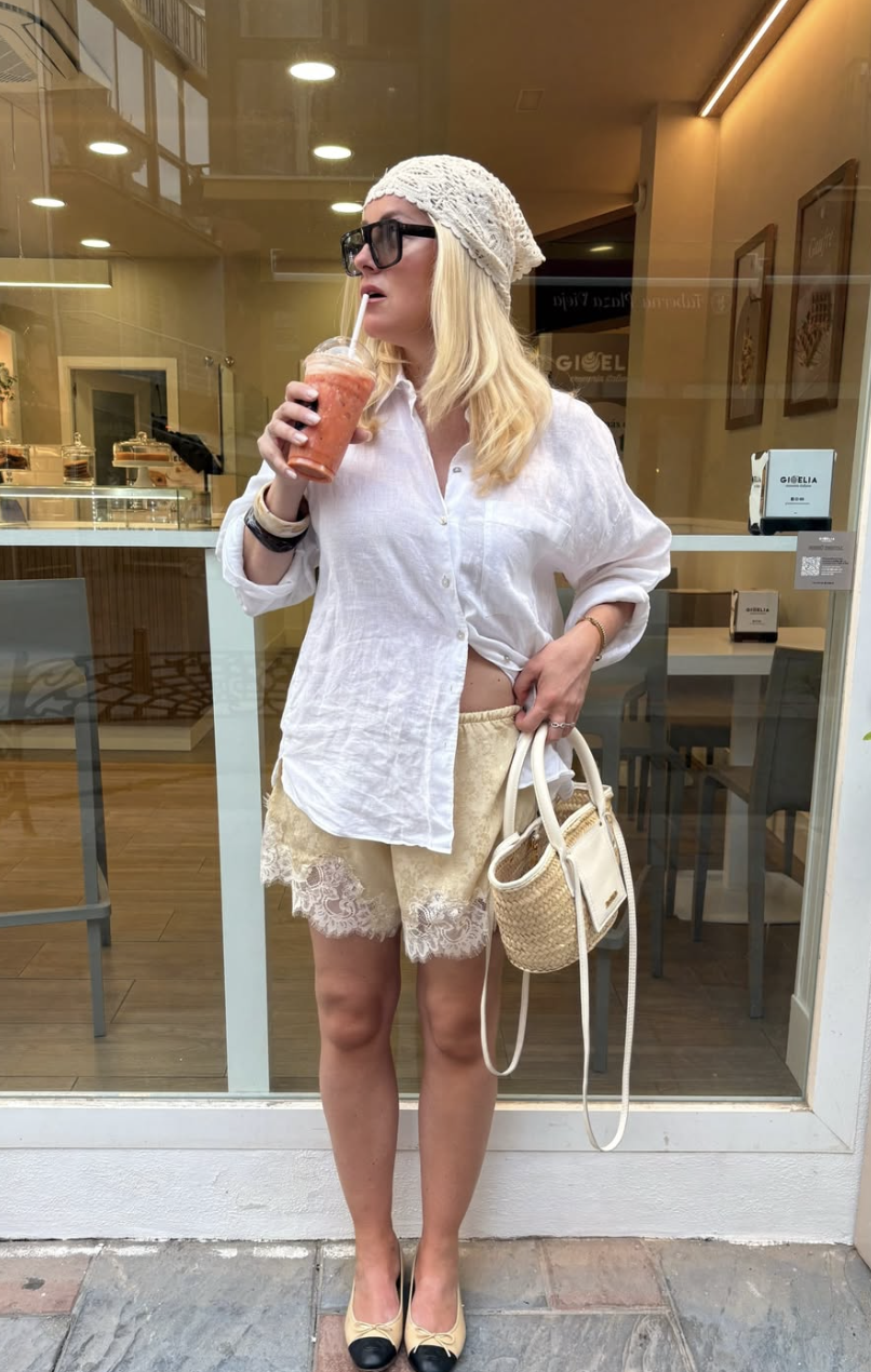 A woman wearing an oversized, unbuttoned white linen shirt over cream-colored lace-trimmed satin shorts, accessorized with a crochet headscarf, sunglasses, and a woven bucket bag.