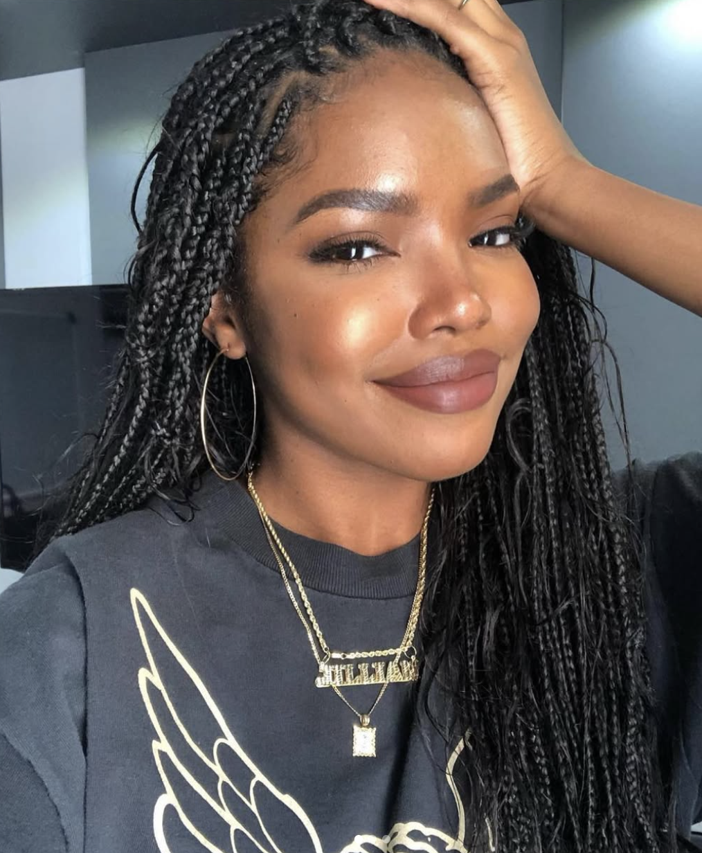 Ryan Destiny showcasing long, black knotless braids with a clean center part, accessorized with large silver hoop earrings and layered gold necklaces.
