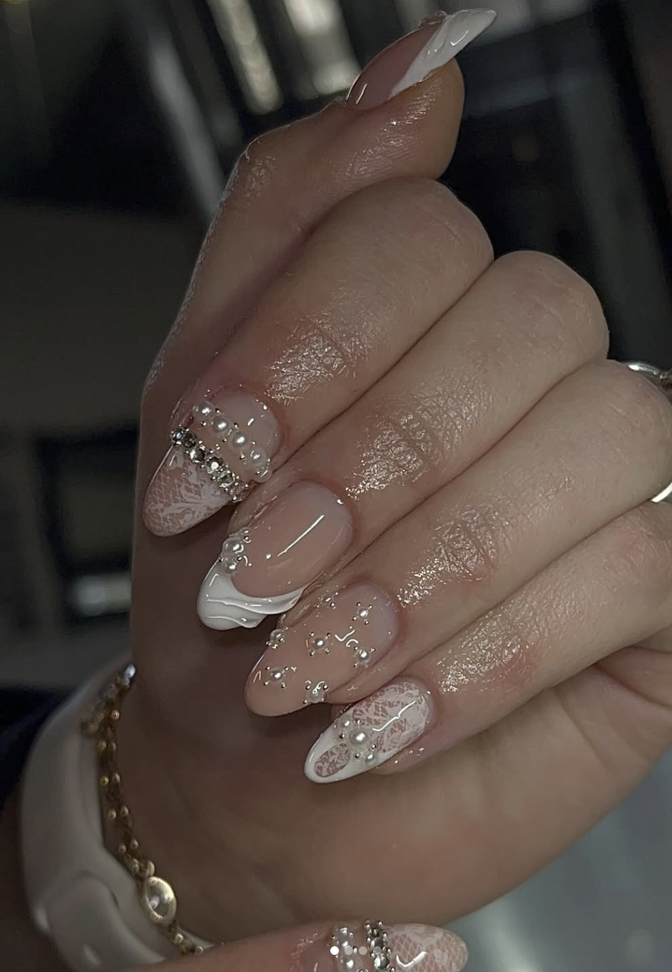 Long almond-shaped nails with a sheer nude base featuring white lace patterns, small pearl accents, and sparkling rhinestone details.