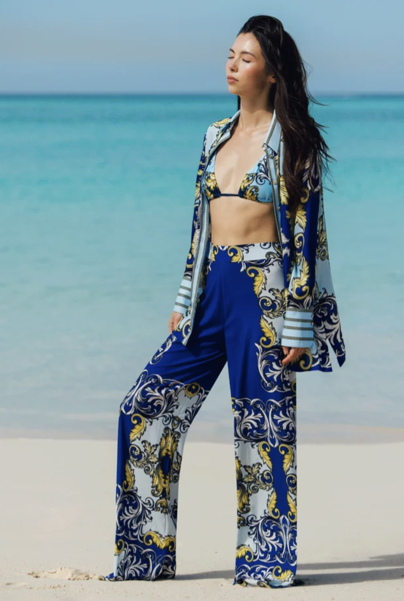 A woman on a beach wearing a blue and white antique scroll print oversized linen button-down shirt, styled open over a matching bikini top with coordinating wide-leg trousers.
