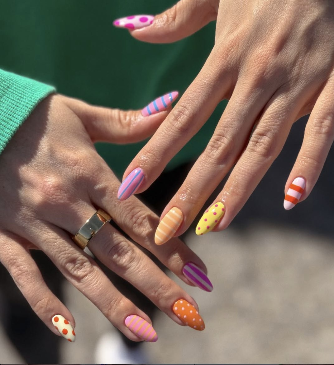 : A close-up of a hand with almond-shaped nails featuring a mismatched "Skittle" design with colorful horizontal stripes and white polka dots in shades of orange, pink, purple, and yellow.