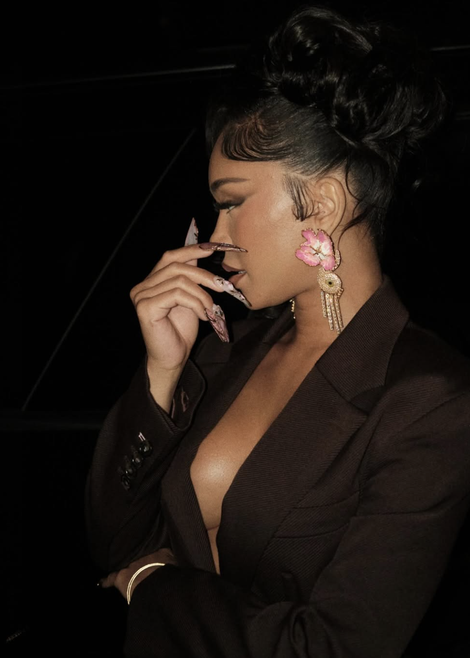 Saweetie wearing a black blazer and large floral earrings, showcasing a half-up high bun with sleek edges to demonstrate how adding height at the crown elongates a round face.