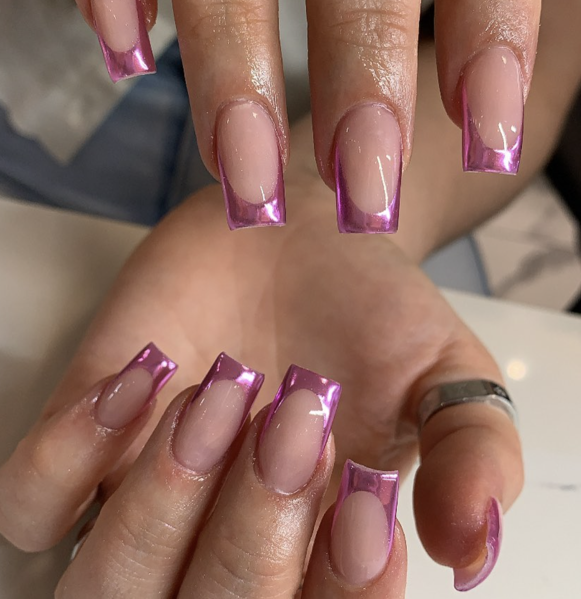 A close-up of medium-length square nails with a sheer neutral base and vibrant pink chrome French tips.