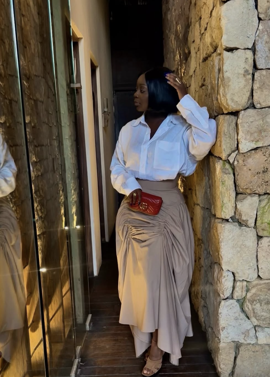 A woman wearing a crisp white button-down shirt tucked into a voluminous, structured taupe maxi skirt with a red clutch bag.