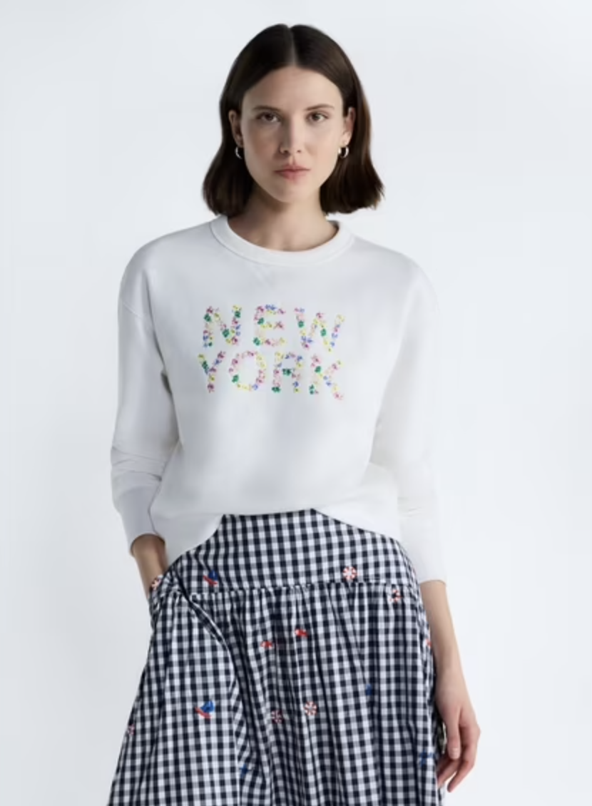 A product shot of a woman wearing a classic white crewneck sweatshirt featuring "NEW YORK" spelled out in a vibrant, multi-colored floral embroidery pattern, styled with black and white gingham trousers.