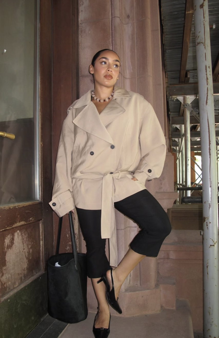A woman wearing a beige trench coat, a statement beaded necklace, and black cropped trousers, carrying a large black leather tote bag.