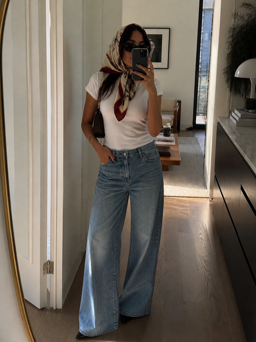 Fashion editor wearing a white t-shirt, wide-leg light wash jeans, and a patterned silk scarf tied over the hair with dark sunglasses.