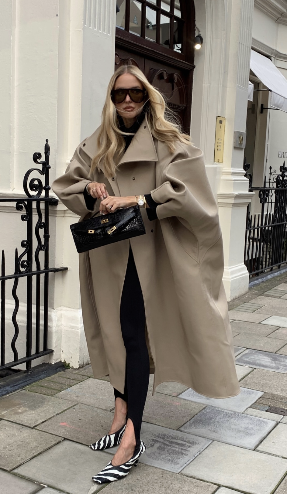 Influencer Leonie Hanne wearing black stirrup leggings paired with zebra-print pointed-toe flats, a long tan trench coat, and carrying a black croc-embossed clutch bag.