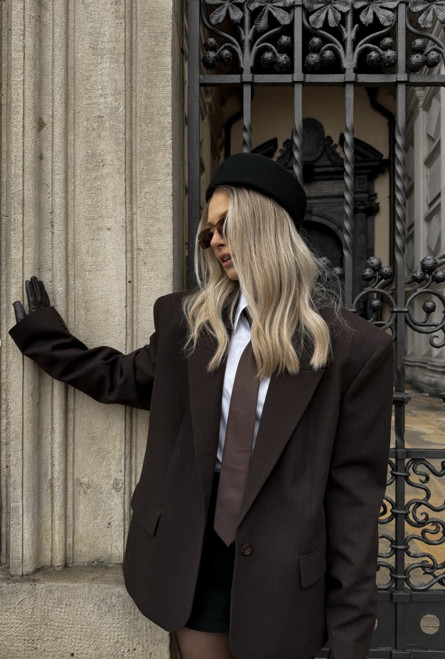 A woman wearing a dark brown structured blazer, a black beret, and a tie, leaning against a stone pillar to showcase the poetcore aesthetic.