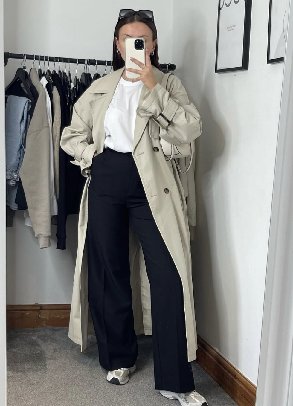 A woman wearing an open cream trench coat layered over a grey hoodie and black wide-leg trousers, styled with white New Balance sneakers for a casual spring outfit.