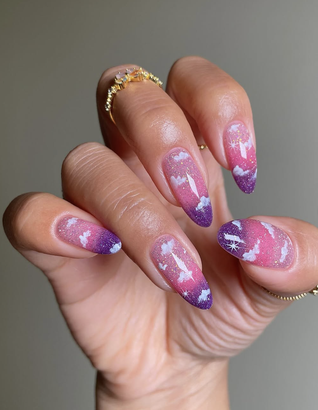 Long almond-shaped gel nails with a pink and purple sunset gradient base, white hand-painted clouds, and subtle glitter.