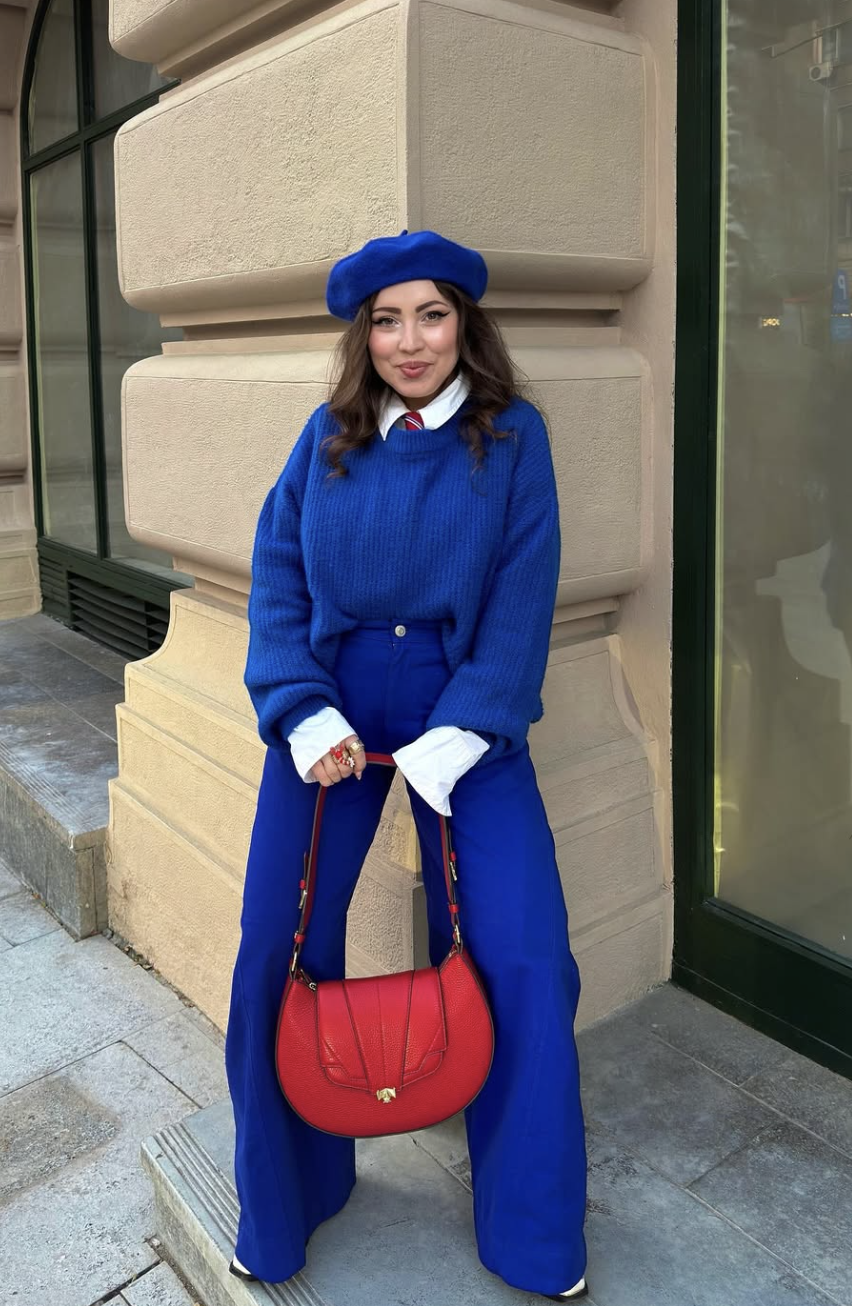 A woman wearing a monochromatic cobalt blue outfit featuring a beret, an oversized knit sweater, and matching trousers with a red handbag.