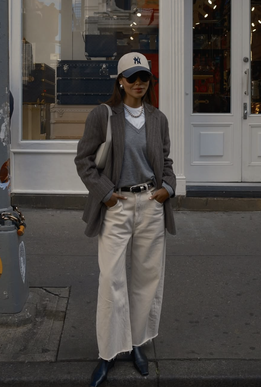 A woman wearing a grey v-neck knit top layered under a grey blazer, styled with white jeans, a New York Yankees baseball cap, and a silver chain necklace.