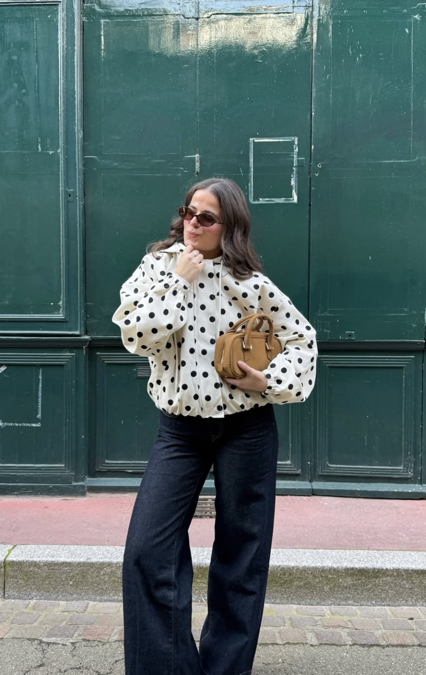 A woman standing in front of a green door wearing an oversized white puffer jacket with large black polka dots, paired with dark-wash wide-leg denim jeans and a tan leather handbag.